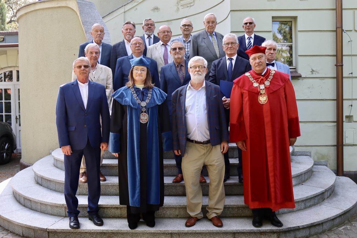 Pamiątkowe zdjęcie grupowe z uroczystości 50-lecia ukończenia studiów. Absolwenci rocznika 1974/1975 Wydziału Melioracji Wodnych pozują na schodach z przedstawicielami władz uczelni, w tym Prorektorem w czerwonej todze i Dziekan w niebieskiej todze.