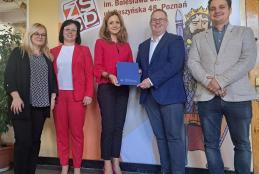 Grupa ludzi (imiona i nazwiska w artykule) stojąca przed ścianą z grafiką. Pięć osób: kobieta w ciemnej bluzce i czarnych spodniach, kobieta w czerwonym garniturze i białej koszuli z ciemnymi włosami i okularami, kobieta w czerwonym garniturze i różowych butach, mężczyzna w granatowym garniturze z niebieską teczką, oraz mężczyzna w jasnej marynarce i ciemnych dżinsach. Tło to historyczna grafika z tekstem, w tym nazwa szkoły.
