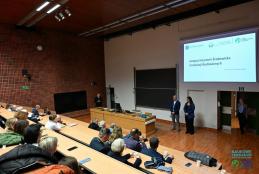 Przestronna aula uniwersytecka z piętrowymi, jasnymi drewnianymi miejscami siedzącymi i dużą grupą osób w różnym wieku. Z przodu prowadzą prezentację Dziekan Wydziału Inżynierii Środowiska i Energetyki Politechniki Poznańskiej profesor Zbigniew Nadolny i profesor Klaudia Borowiak Dziekan Wydziału Inżynierii Środowiska i Inżynierii Mechanicznej na tle ekranu z polskim tekstem o inteligencji instytucjonalnej w budownictwie. Aula ma ściany z czerwonej cegły, jasny sufit. Kilka osób stoi przy wyjściu.