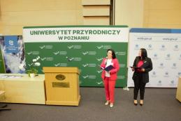 Dwie kobiety na podium, przemawiające do publiczności, za nimi jest zielony ekran z napisem - Uniwersytet Przyrodniczy w Poznaniu