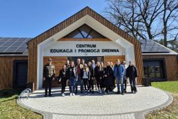 Zdjęcie grupowe studentów drugiego stopnia, II roku Gospodarki Przestrzennej z prof. UPP dr. hab. inż. Adamem Zydroniem oraz dr. inż. Cyprianem Chwiałkowskim przed budynkiem z panelami słonecznymi. Na budynku widoczny napis "Centrum Edukacji i Promocji Drewna"