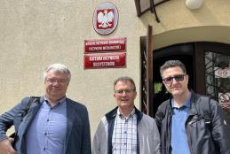 Od lewej Profesor Shkumbin Makolli - włosy koloru siwego okularach i granatowej marynarce.  Doktor inżynier Mirosław Czechlowski - w okularach i szarej koszuli w kratę i profesor Ruslan Mariychuk - w ciemnej kurtce i okularach stoją przed jasnobeżowym budynkiem. Na fasadzie budynku widoczne prostokątne tablice. Na jednej z tablicy nazwa wydziału, na drugiej tablicy napis "Katedra Inżynierii Biosystemów". Nad prostokątnymi tablicami, owalna tabliczka godłem Polski. Jasne światło sugeruje słoneczny dzień.