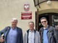 Od lewej Profesor Shkumbin Makolli - włosy koloru siwego okularach i granatowej marynarce.  Doktor inżynier Mirosław Czechlowski - w okularach i szarej koszuli w kratę i profesor Ruslan Mariychuk - w ciemnej kurtce i okularach stoją przed jasnobeżowym budynkiem. Na fasadzie budynku widoczne prostokątne tablice. Na jednej z tablicy nazwa wydziału, na drugiej tablicy napis "Katedra Inżynierii Biosystemów". Nad prostokątnymi tablicami, owalna tabliczka godłem Polski. Jasne światło sugeruje słoneczny dzień.