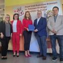 Grupa ludzi (imiona i nazwiska w artykule) stojąca przed ścianą z grafiką. Pięć osób: kobieta w ciemnej bluzce i czarnych spodniach, kobieta w czerwonym garniturze i białej koszuli z ciemnymi włosami i okularami, kobieta w czerwonym garniturze i różowych butach, mężczyzna w granatowym garniturze z niebieską teczką, oraz mężczyzna w jasnej marynarce i ciemnych dżinsach. Tło to historyczna grafika z tekstem, w tym nazwa szkoły.