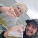 Kobieta i mężczyzna przy zbiorniku wodnym. Kobieta ma jasne włosy, uśmiecha się. Mężczyzna w ciemnej kurtce z kapturem, uśmiecha się i patrzy w obiektyw aparaty fotograficznego. Oboje trzymają przezroczyste woreczki z materiałem badawczym. Tło to jasnoszare, pochmurne niebo.