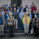 Grupa ludzi stojąca na schodach (uczestnicy konferencji). Osoby w różnym wieku i płci, ubrani w casualowe ubrania (dżinsy, kurtki, swetry). Jedna kobieta w centralnej części ma pasiaste, musztardowo-żółte i brązowe ubranie. Tło sugeruje zewnętrzne wejście lub patio z jasną ścianą.