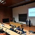 Przestronna aula uniwersytecka z piętrowymi, jasnymi drewnianymi miejscami siedzącymi i dużą grupą osób w różnym wieku. Z przodu prowadzą prezentację Dziekan Wydziału Inżynierii Środowiska i Energetyki Politechniki Poznańskiej profesor Zbigniew Nadolny i profesor Klaudia Borowiak Dziekan Wydziału Inżynierii Środowiska i Inżynierii Mechanicznej na tle ekranu z polskim tekstem o inteligencji instytucjonalnej w budownictwie. Aula ma ściany z czerwonej cegły, jasny sufit. Kilka osób stoi przy wyjściu.