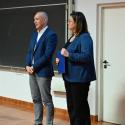 Profesor Zbigniew Nadolny i profesor Klaudia Borowiak podczas prelekcji stoją na jasnej drewnopodobnej podłodze. Profesor Nadolny, po lewej, jest łysy, ubrany w granatową marynarkę i jasnoszare spodnie, z rękami opartymi na biodrach, patrzy przed siebie. Profesor Borowiak, po prawej, ma brązowe włosy do ramion, nosi granatową marynarkę i ciemne spodnie, trzyma dokument i patrzy lekko w lewo. W tle tablice kredowe, biała ściana i częściowo widoczne białe drzwi.