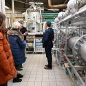 Grupa osób odwiedzających laboratorium lub obiekt przemysłowy. Na pierwszym planie duże metalowe urządzenie przemysłowe, prawdopodobnie wymiennik ciepła, z licznymi połączeniami śrubowymi i rurami. Wokół niego stoi kilku mężczyzn i dwie kobiety. Jedna z kobiet, w pomarańczowym płaszczu, wyróżnia się na pierwszym planie. W tle widać inne urządzenia, w tym metalową jednostkę przetwórczą. Wnętrze laboratorium ma jasną podłogę i neutralne kolory. Oświetlenie wskazuje na dobrze oświetlone środowisko.