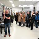 Uczestnicy seminarium w laboratorium. Na pierwszym planie jasna podłoga, grupa stoi w półkolu, zwrócona w stronę mężczyzny w czarnej koszulce i niebieskich dżinsach, który gestykuluje. Większość osób ma na sobie płaszcze, kurtki lub kardigany w neutralnych kolorach. W tle widoczny nowoczesny wystrój laboratorium z metalowymi klatkami i urządzeniami na stole. W prawym górnym rogu osoba w ciemnej kurtce, obserwująca grupę.