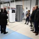 Uczestnicy seminarium w laboratorium, którzy oglądają biały cylindryczny podgrzewacz wody lub zbiornik solarny.  W tle jasne, dobrze oświetlone pomieszczenie techniczne. Widoczny mężczyzna z krótkimi brązowymi włosami w kurtce, mężczyzna w garniturze oraz młoda kobieta w jasnym stroju.