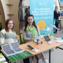 Dwie studentki Studenckiego Koła Naukowego Ekoenergetyki siedzą przy swoim stoisku z panelami słonecznymi, ulotkami i miniaturowym pojazdem solarnym