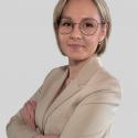 Doktor inżynier Patrycja Mleczek w jasnobeżowej marynarce i naszyjniku, stojąca pewnie z założonymi rękami na tle neutralnej, szarej ściany.