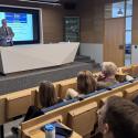 Prezentacja odbywa się w nowoczesnej, jasnej sali konferencyjnej. Mężczyzna w garniturze stoi przy podwyższonej, jasnoszarej mównicy, zwrócony w stronę dużego ekranu z akademicką prezentacją. Publiczność siedzi na jasnobrązowych, modułowych siedziskach, wielu uczestników jest widocznych częściowo od tyłu i skupionych na prezentacji. Ściany mają jasny, drewniany odcień, a podłoga jest pokryta szarą wykładziną.