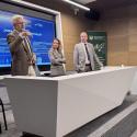 Prezentacja odbywa się w nowoczesnej sali konferencyjnej. Mężczyzna w jasnobrązowo-szarej marynarce i jasnych spodniach gestykuluje podczas przemówienia przy długim, białym stole prezentacyjnym. Obok stoi kobieta w podobnej marynarce i jasnych spodniach. Za stołem stoi mężczyzna w ciemnoszarym garniturze lub marynarce. W pomieszczeniu dominują jasne ściany i sufit, podłoga jest pokryta szarą wykładziną, a w pobliżu stołu ustawione są szare krzesła. 