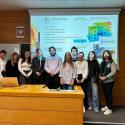 Dwóch wykładowców  i studenci - stoją w sali wykładowej przed dużym ekranem prezentacyjnym. Na ekranie widoczne są wykresy, diagramy techniczne i inne treści związane prawdopodobnie z inżynierią konstrukcyjną. Część osób ma na twarzach nałożone projekcje zdjęć innych ludzi, będące częścią prezentacji. Przed grupą znajduje się drewniany stół, na którym stoi laptop. Ściany sali są wyłożone jasnobrązowymi panelami drewnianymi.