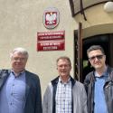 Od lewej Profesor Shkumbin Makolli - włosy koloru siwego okularach i granatowej marynarce.  Doktor inżynier Mirosław Czechlowski - w okularach i szarej koszuli w kratę i profesor Ruslan Mariychuk - w ciemnej kurtce i okularach stoją przed jasnobeżowym budynkiem. Na fasadzie budynku widoczne prostokątne tablice. Na jednej z tablicy nazwa wydziału, na drugiej tablicy napis "Katedra Inżynierii Biosystemów". Nad prostokątnymi tablicami, owalna tabliczka godłem Polski. Jasne światło sugeruje słoneczny dzień.