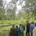 Grupa osób stoi na zielonym brzegu strumienia Bogdanka, otoczona drzewami i bujną roślinnością. Patrzą w kierunku strumienia i dalej w gęsto zalesiony teren, podczas spaceru edukacyjnego poświęconego renaturyzacji cieków miejskich.